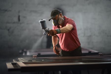 Front view of senior man in red T-shirt and protective mask and gloves painting fence with polyvizor in brown colour in smithy. Concept of process painting fence.